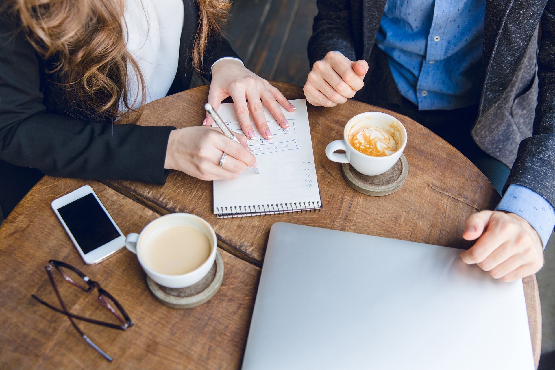 Två kollegor antecknar vid ett runt träbord med kaffekoppar och en mobiltelefon